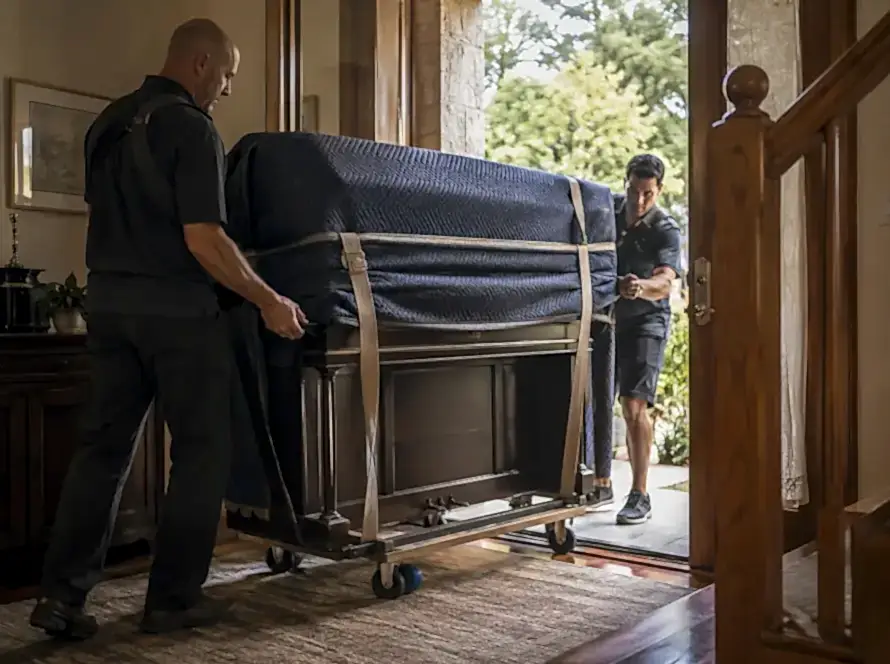 Professional piano movers carefully transporting an upright piano through a residential doorway using a dolly and protective moving blankets.
